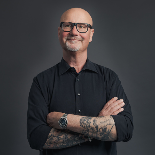 frieder barth, ceo von friederbarth, porträtfoto mit verschränkten armen, tätowierten unterarmen und schwarzem hemd vor dunklem hintergrund