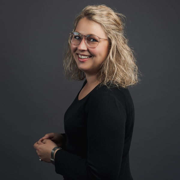svea bielert, ceo und projektmanagerin bei friederbarth, porträtfoto mit blonden, gewellten haaren und schwarzem oberteil vor dunkelgrauem hintergrund