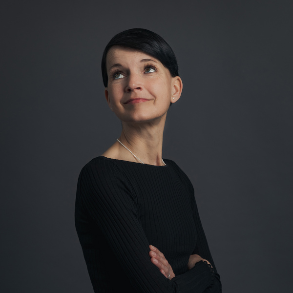stefanie brandt, premium professional bei friederbarth, porträtfoto mit kurzem dunklem haar, schwarzem oberteil und halskette vor dunklem hintergrund