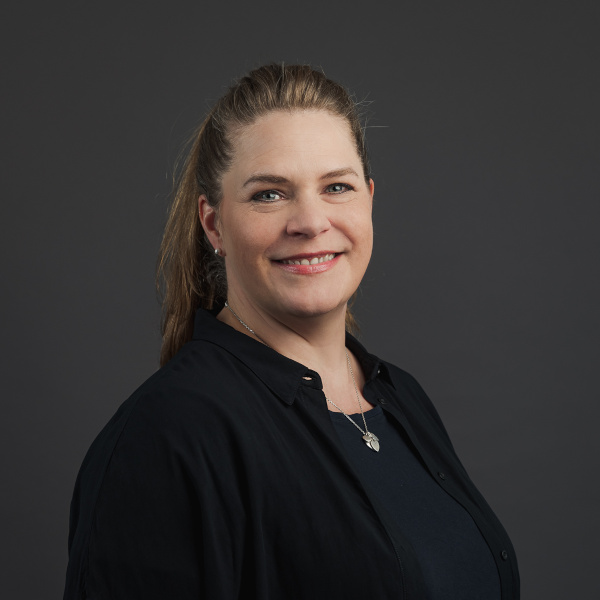 anke dahnke, management- und team-assistentin bei friederbarth, porträtfoto mit langen gewellten haaren und herzförmiger halskette vor dunkelgrauem hintergrund