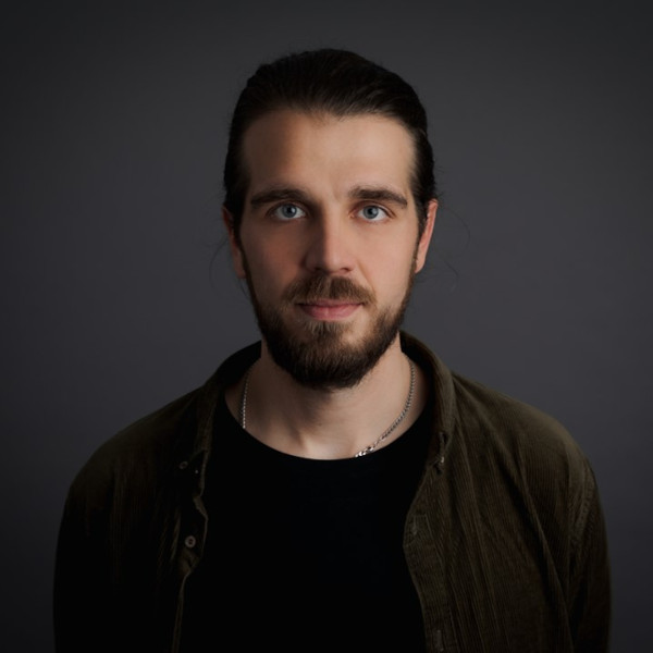 Adrian Herrmann mit zurückgebundenem Haar, dunklem Shirt und offener Hemdbluse – Portraitaufnahme vor dunklem Hintergrund mit unscharfem Gesicht