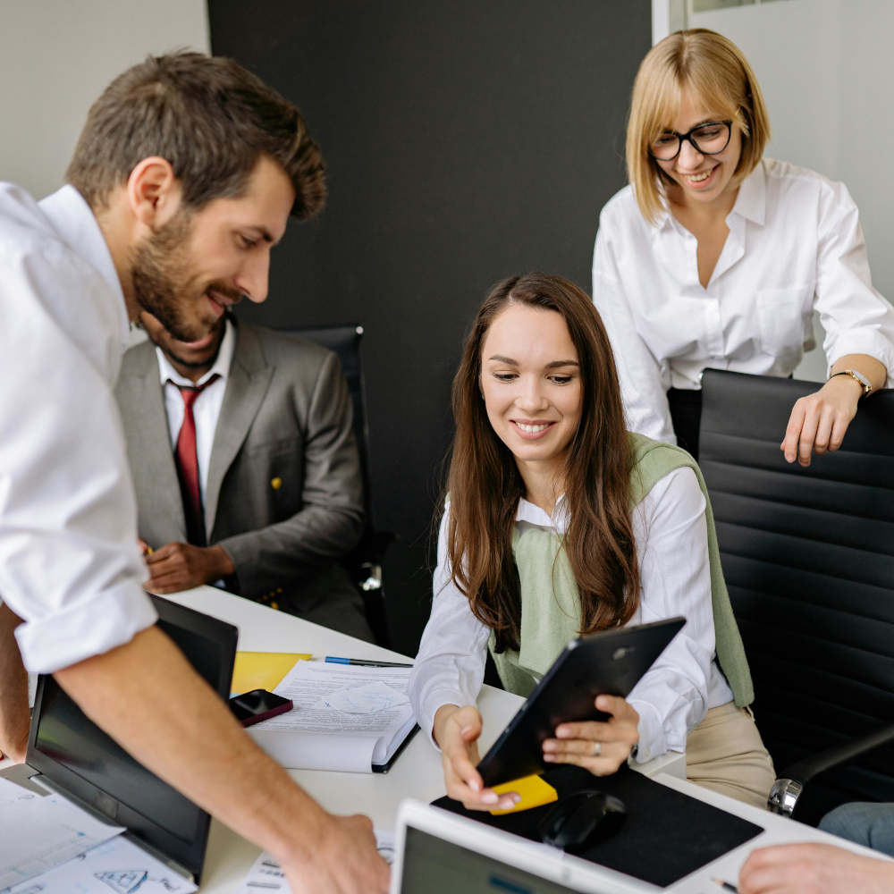 Fünf Personen in formeller Kleidung sitzen und stehen um einen Konferenztisch in einem modernen Büro. Sie diskutieren über Dokumente, Laptops und Tablets – eine Szene, die professionelle Teamarbeit und Führungskompetenz vermittelt.