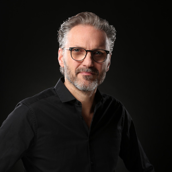 Guido Körfer in schwarzem Shirt vor dunklem Hintergrund – Portraitaufnahme mit unscharfem Gesicht