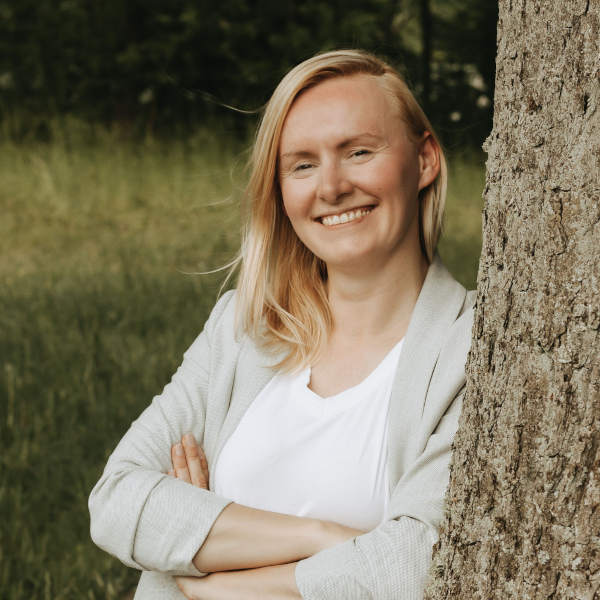 Mareen Mallon mit weißem Shirt, grauem Cardigan und dunkler Jeans, stehend mit verschränkten Armen an einem Baum im Grünen – Gesicht unscharf