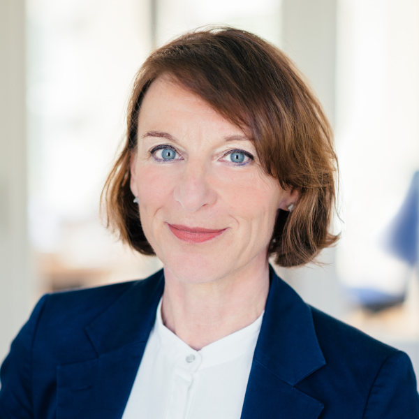 Martina Knobloch mit kurzem, hellbraunem Haar im dunkelblauen Blazer und weißem Shirt – Portraitaufnahme mit unscharfem Gesicht in heller Innenraumszene