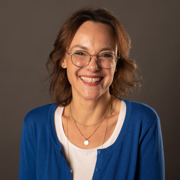 Melanie Wilden im blauen Cardigan über weißem Shirt mit zwei Halsketten – Portraitaufnahme mit unscharfem Gesicht vor dunklem Hintergrund