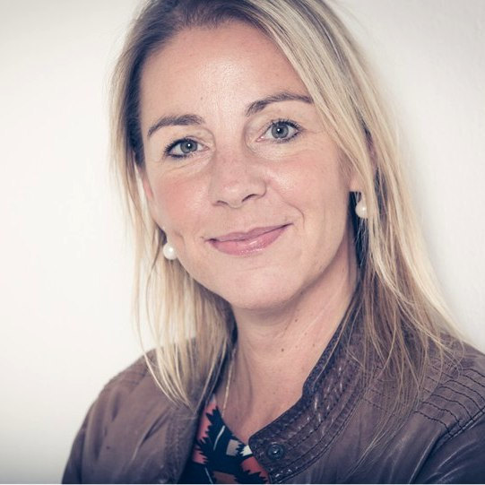 Sabine Fries mit langen blonden Haaren in brauner Lederjacke und gemustertem Shirt – Portraitaufnahme mit unscharfem Gesicht