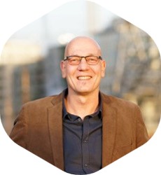 Person mit Brille und brauner Jacke vor urbanem Hintergrund – symbolisch für professionelle, vertrauensvolle Zusammenarbeit im Kommunikationstraining mit FRIEDERBARTH.