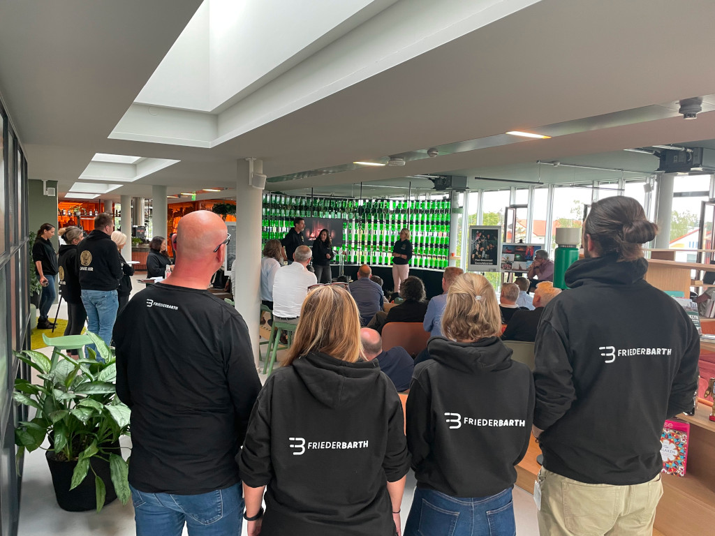 Teilnehmende eines Mitarbeiterworkshops bei Mast Jägermeister in einem modernen Raum mit großen Fenstern und grünen Jägermeister-Flaschen im Hintergrund – alle tragen schwarze Hoodies mit „FRIEDERBARTH“-Aufdruck.